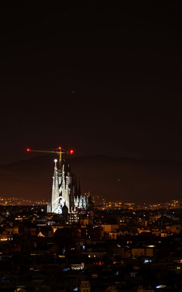 Die beleuchtete Sagrada Família ragt bei Nacht aus den dunklen Dächern des Eixample, dahinter die Hügelkette