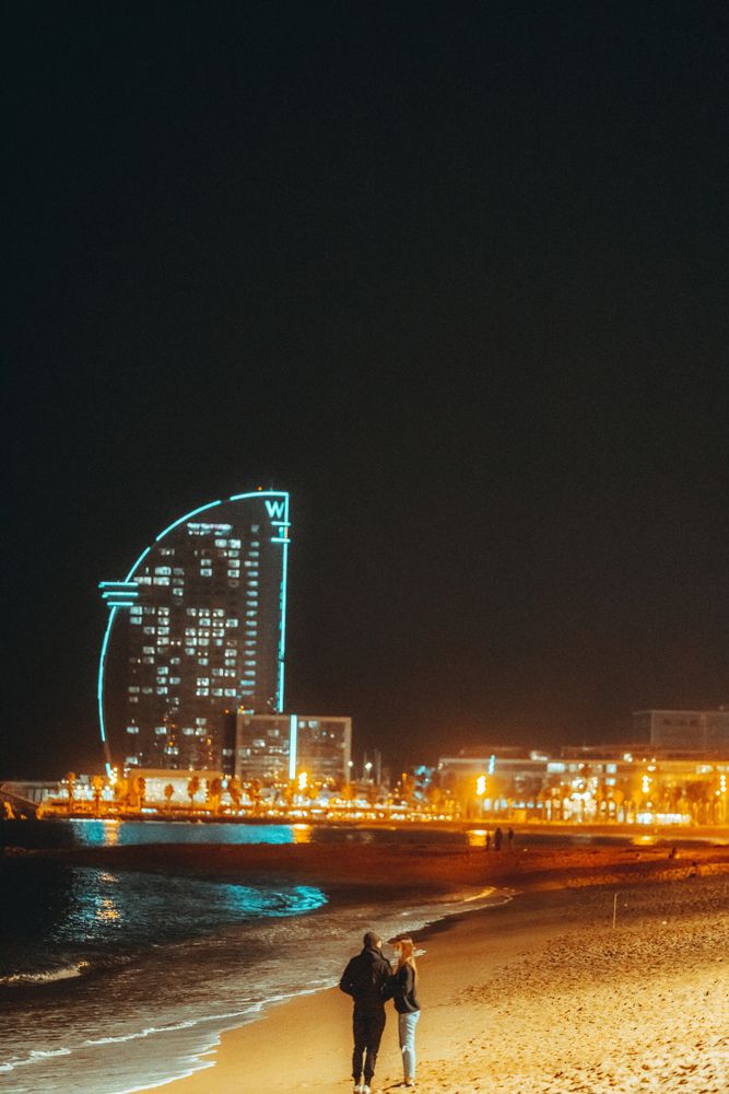 Strand der Barceloneta bei Nacht mit dem beleuchteten segelförmigen Hotel und zwei Personen am Wasser